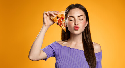 Girl shows slice of delicious pizza, makes kissing face from satisfaction. Woman eating in favorite pizzeria restaurant, orange background