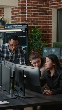 Vertical video Software engineers brainstorming ideas looking at code on computer screens asking feedback from senior developer while intern joins the discussion. Junior programmers collaborating on