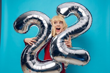 a fervently laughing, emotional woman in a red shirt stands on a blue background and holds inflatable balloons in the shape of the number twenty-two in silver color, hugging them with her hands