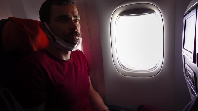 Man has pulled down medical mask on chin during the flight on plane and then worn it back when somebody asked him. A young bearded tourist travelling by jet looks through the window with serious face.