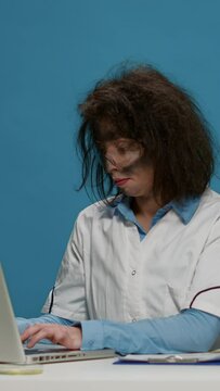 Vertical video: Portrait of crazy female chemist working on laptop at desk, being funny and goofy in studio. Mad insane scientist using computer and doing foolish silly facial expressions, having