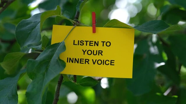 A paper note with the words Listen To Your Inner Voice  on it attached to a tree with a clothes pin. Close up.