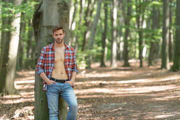 Handsome unshaven man with lumbersexual look in unbuttoned lumberjack shirt forest background