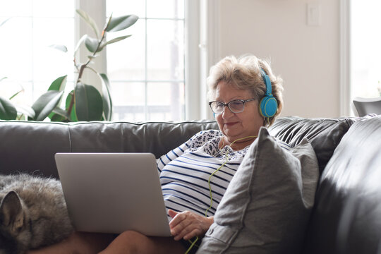 Senior woman in headphones sitting on couch at home using laptop computer
