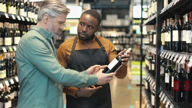 African american male seller in apron talking with handsome man while standing between rows of shelves with bottles. Shop assistant recommending wine to customer in alcohol store department.
