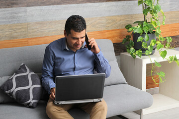 Single Latino adult man does home office in his living room with laptop and cell phone, creates...