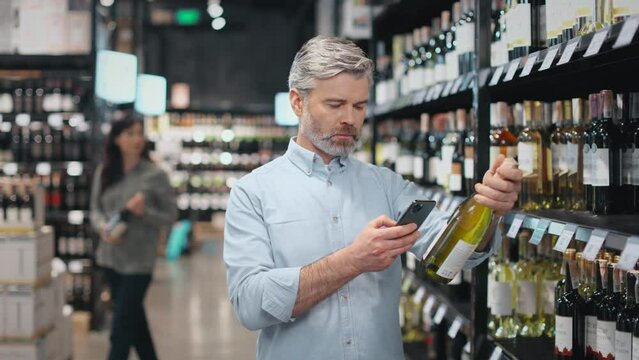 Caucasian aged man in casual attire searching information about wine in internet using smartphone. Male customer choosing and buying alcohol at store with help of modern gadget.