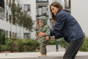 Fototapeta premium A mother is holding her pretty daughter in a raincoat on a cloudy day. A mom is teaching her female toddler how to do steps in the street at noon.