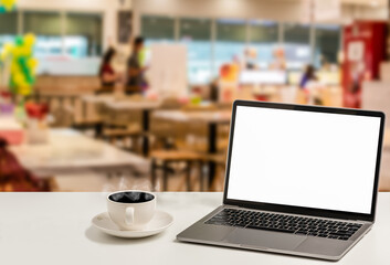 A blank white screen laptop and coffee cup are placed on a white desk in a food court. The Concept for business, technology, internet, design, and art. Closeup, selective focus, blurred background