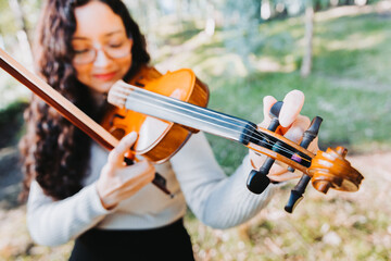 Curly brunette woman with glasses tuning her violin outside in the woods. Selective focus.