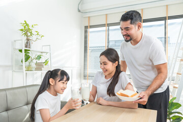 Asian  family enjoying breakfast at living room. little girl daughter sitting on table, drinking milk with smiling father and mother in morning. Happy family at home.