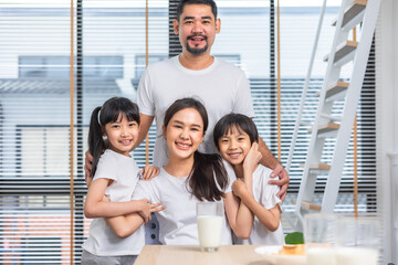 Obraz premium Asian family enjoying breakfast at living room. little girl daughter sitting on table, drinking milk with smiling father and mother in morning. Happy family at home.