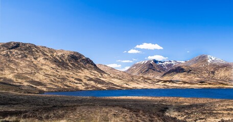 Beautiful calm loch or lake surrounded by rugged hills, brown scrub grassland and blue sky with a few clouds. Copy space. Scottish Highlands, Scotland, UK.