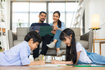 Happy Asian family relaxing on sofa while kid drawing on floor. Little boy girl having fun, friendship between siblings, family leisure time in living room at home.