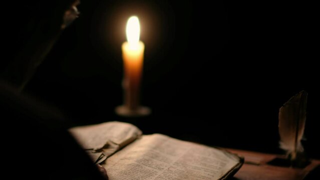 Video of old monk reading old book in dark monastic cell room under candle light.