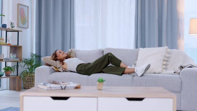 Smiling woman relaxing on sofa in home living room as new property homeowner and real estate investor. Relieved, successful and accomplished lady lying with hands behind her head in a purchased house