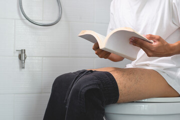 A young man wearing white t-shirt with diarrhea sits at home reading a book on the toilet. He had a...