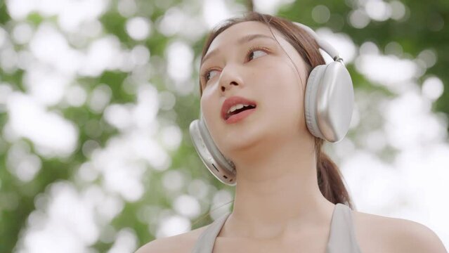 Asian woman is engaged in fitness in public park. Female wearing sport bra and using headphone listening to music. Healthy lifestyle concept, sports outdoor activities in park