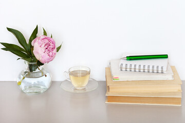 A stack of old books with bookmarks, a notepad with a pen for notes, evening herbal tea in a glass cup and a pink peony flower on a light table. Home comfort, spring still life