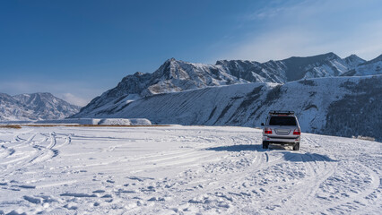 Fototapeta na wymiar The car is parked on a snow-covered high-altitude plateau. Footprints and tire tracks in the snow. A picturesque mountain range against the blue sky. Altai