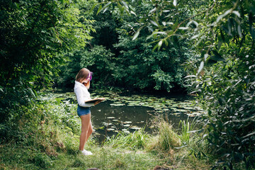 Naklejka na ściany i meble A young schoolgirl with stylishly dyed hair, with a purple lock of hair, in denim shorts and a white shirt, stands by a picturesque river in the forest and reads a book.