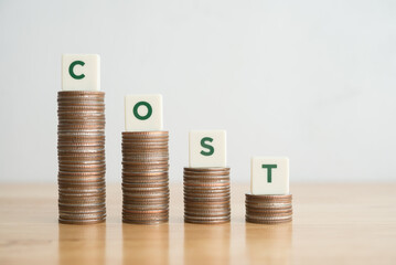 Word COST on stacks coin as graph chart falling down on wooden table background. Concept of cost reduction, cost cut in business company or production, optimize continuous improvement.