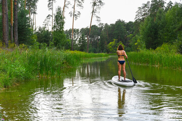 A woman drives on the Sup Board through a narrow canal surrounded by dense grass. Active weekend vacations wild nature outdoor. The woman is standing in a bathing suit.