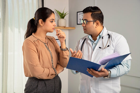 Indian man or male physician or doctor wearing stethoscope and apron holding a report file in hand and giving consultation to a tensed or worried female or Woman patient. Medical, medicine, healthcare