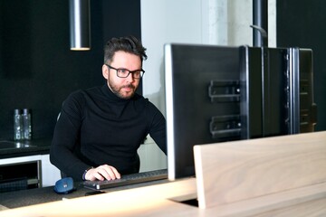 Portrait of handsome focused concentrated young bearded man is using pc computer at home, luxury apartment in glasses. Freelancer is working, looking at screen. Distant freelance job, work.
