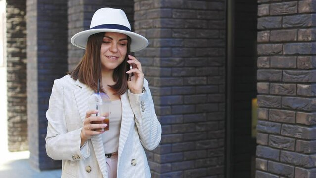 Beautiful woman going to meeting with cold coffee and talking to the phone walking on the street. Portrait of successful business woman holding cup of cold drink in hand on her way to work