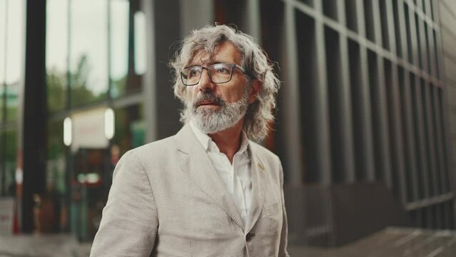 Mature businessman with beard in eyeglasses wearing gray jacket walks down the street past modern buildings