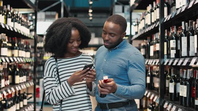 African american couple dressed in casual attire standing at wine store and making photo of bottle label on smartphone. Married young couple smiling, talking and looking on each other.