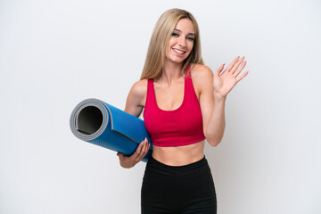 Young sport blonde woman going to yoga classes while holding a mat isolated on white background saluting with hand with happy expression