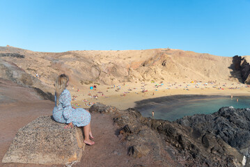 una turista con pelo rubio y vestido azul sentada en una roca mirando la playa de Papagayo en Lanzarote en las Islas Canarias. Relax en verano en la costa. 