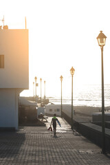 Ni&ntilde;o surfista de espaldas paseando por las calles de Caleta caballo, con un neopreno negro y amarillo y sus aletas y la tabla de surf en las manos durante el atardecer en Lanzarote, Islas Canarias