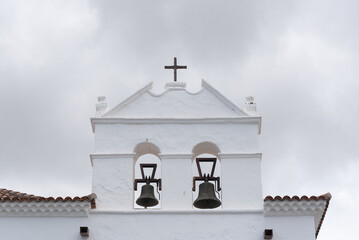 Plano detalle del campanario de una iglesia, la fachada que alberga dos campanas y es de color blanco, termina en un tímpano culminado por una cruz de hierro, de fondo un cielo blanco nuboso.