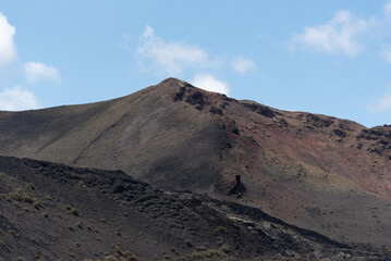 Paisaje volcánico y desértico con tierra negra y rocas oscuras y cos volcanes inactivos durante un día de verano en Lanzarote, Islas Canarias. Recursos turísticos y naturales.