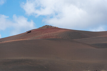 Fototapeta na wymiar Detalle de un paisaje desértico y volcánico de tierras en tonos marrones y naranjas con un gran volcán inactivo al fondo durante un día nublado de verano en Lanzarote, Islas Canarias.