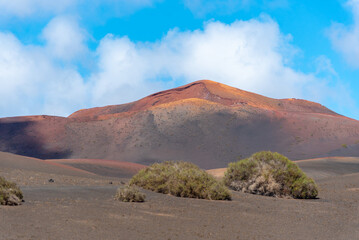Paisaje desértico y volcánico con un gran volcán inactivo al fondo y algo de vegetación en primer plano  durante un día soleado de verano en Lanzarote, Islas Canarias.
