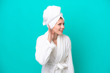 Young woman in a bathrobe isolated on blue background listening to something by putting hand on the ear