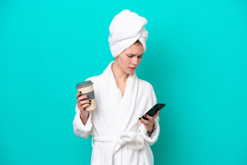 Young woman in a bathrobe isolated on blue background holding coffee to take away and a mobile