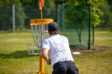 Caucasian male throwing a disc golf to the basket outdoors on a sunny day. He is wearing a black cap. Sunny sports day in the park. - 518741434