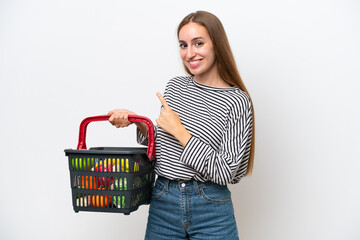 Young Rumanian woman holding a shopping basket full of food isolated on white background pointing finger to the side