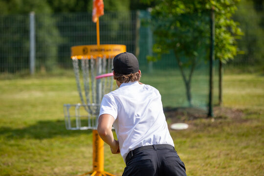 Caucasian male throwing a disc golf to the basket outdoors on a sunny day. He is wearing a black cap. Sunny sports day in the park.