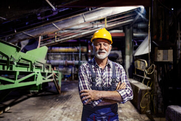 A happy senior factory worker stands in the dark with arms crossed and looks at the camera.
