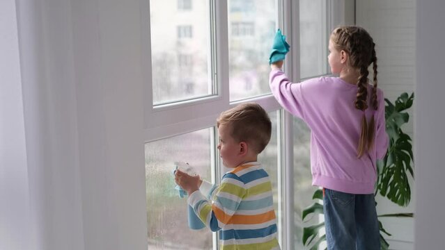 A little boy and girl splashes water from a bottle on the window and wipes it with a rag. Children help with cleaning the house. Clear the window. Brother and sister cleaning the window.