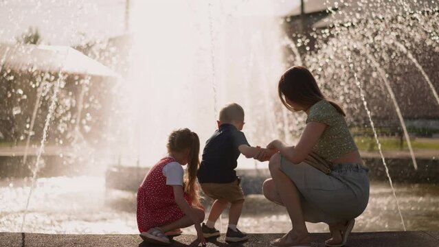 Mother with little son and daughter sits on parapet surrounding large fountain in park backside view slow motion. Woman and children refresh with splashing water drops