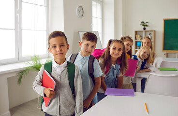 Fototapeta premium Education for children. Cheerful cute elementary school students pose in classroom on first of September. Little boys and girls with books and backpacks stand one behind other and smile at camera.