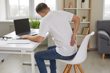 Man experiences severe back pain caused by prolonged and incorrect sitting at workplace. Young man sitting with his back to camera holds his lower back, feeling exhausted from sedentary lifestyle.