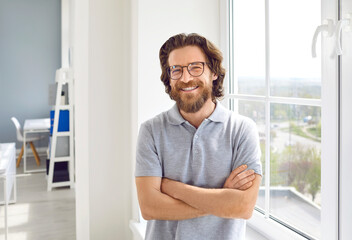 Portrait of a happy successful businessman. Cheerful smiling handsome bearded young Caucasian business man in a casual grey polo shirt and glasses standing with his arms crossed by the office window